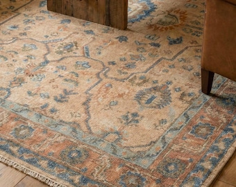 Op de afbeelding: Een beige vloerkleed met een gedetailleerd bloemenpatroon in blauwe, oranje en groene tinten. Het kleed heeft een verweerde uitstraling en een gefranjerde rand. Het ligt op een houten vloer, met een houten tafel en leren stoel in beeld.