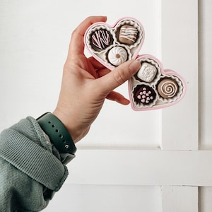 May include: A pink heart-shaped box of chocolates, held in a hand. The box contains various chocolates with different designs and colors. The chocolates are in a white paper cup. The box is held against a white background.