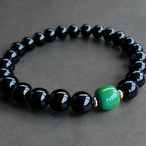 Puede incluir: Una pulsera de cuentas negras con una sola cuenta verde y detalles dorados. La pulsera presenta cuentas negras redondas y pulidas, con una vibrante cuenta verde en el centro.