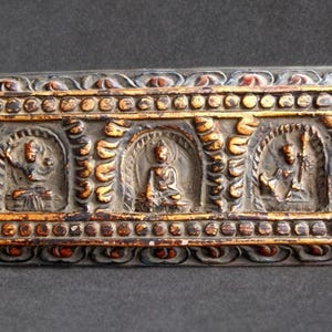 Puede incluir: Pieza decorativa rectangular de estilo antiguo con tres figuras talladas en nichos arqueados. La pieza tiene una base gris plateado con detalles dorados y marrones, y un borde detallado. Las figuras parecen estar en una postura sentada o meditativa.
