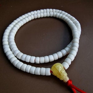 Puede incluir: Collar de cuentas blancas con un colgante amarillo y una cuerda roja. El collar está hecho de pequeñas cuentas cuadradas. El colgante es de color amarillo claro y tiene una forma única. La cuerda roja está atada en la parte inferior del colgante.