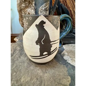 Puede incluir: Una taza de cerámica con un diseño en blanco y negro de un oso esquiador. La taza tiene un asa azul y detalles de copos de nieve. La taza está sobre una superficie de piedra gris.