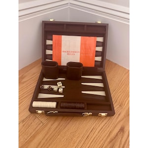 May include: A brown backgammon set in a case with white and brown triangular points, two dice cups, four dice, and a stack of white and brown checkers. An orange booklet labeled BACKGAMMON RULES is placed inside the open game board.