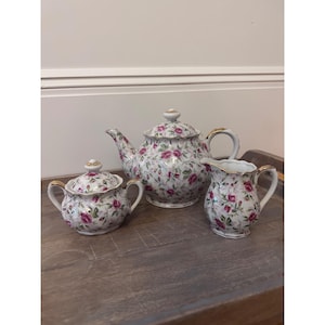 Op de afbeelding: Een wit porseleinen theeservies bestaande uit een theepot, een suikerpot met deksel en een melkkannetje, versierd met een patroon van kleine roze roosjes en groene blaadjes, afgewerkt met gouden randen op de handvatten en deksels.
