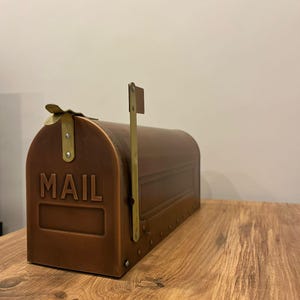 Puede incluir: Un buzón de color cobre con la palabra "MAIL" en relieve en la parte delantera. El buzón tiene una parte superior curvada, una bandera y detalles en color latón. El buzón está sobre una superficie de madera.