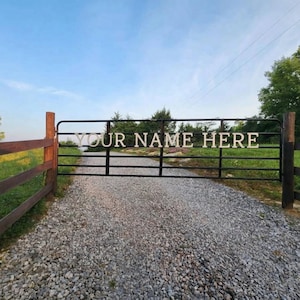 Pode incluir: Um portão de metal preto com o texto "YOUR NAME HERE" em letras brancas. O portão está situado entre postes de madeira e uma entrada de cascalho, com um céu azul e árvores ao fundo.