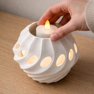 Può includere: Un portacandele in ceramica bianca con un design a spirale e ritagli ovali. All'interno è posizionata una candela bianca con una luce gialla tremolante. Il portacandele è appoggiato su una superficie di legno, creando un'atmosfera calda.