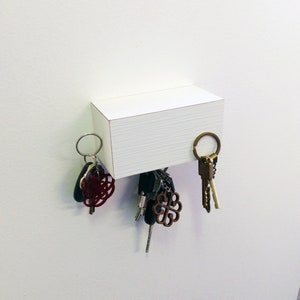 May include: White magnetic key holder with a wood grain texture.  Keys are hanging on the holder.  The keys are silver and gold with a red and black key chain.