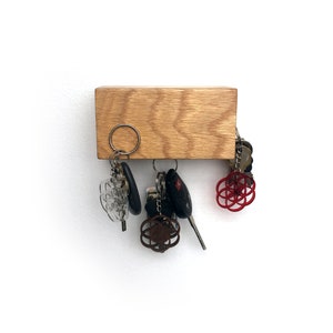 May include: A wooden key holder with a natural wood finish. Five keys are hanging from the holder, including a car key with a black fob, a clear plastic key with a silver ring, and a silver key with a red and silver charm. The key holder is mounted on a white wall.