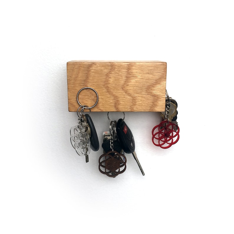 May include: A wooden key holder with a natural wood finish. Five keys are hanging from the holder, including a car key with a black fob, a clear plastic key with a silver ring, and a silver key with a red and silver charm. The key holder is mounted on a white wall.