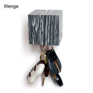 May include: A wooden key holder with a black and white wood grain pattern. The holder is shaped like a cube and has a metal hook for hanging keys. The word "Wenge" is written above the holder.