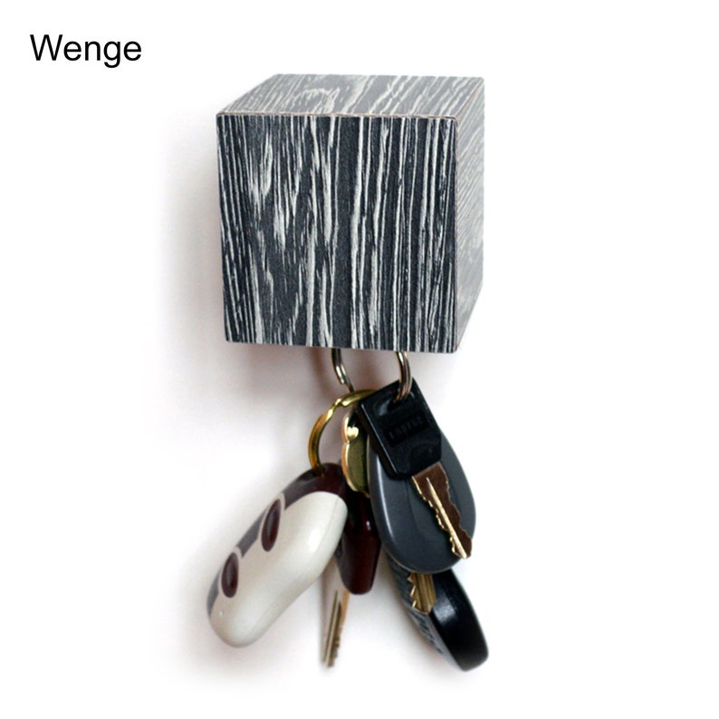 May include: A wooden key holder with a black and white wood grain pattern. The holder is shaped like a cube and has a metal hook for hanging keys. The word "Wenge" is written above the holder.
