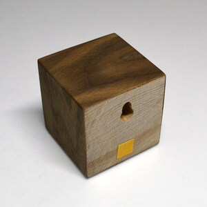 May include: A small wooden cube with a hole in the center and a yellow adhesive square on the bottom. The cube is made of dark wood with a light wood grain.