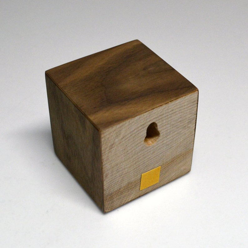 May include: A small wooden cube with a hole in the center and a yellow adhesive square on the bottom. The cube is made of dark wood with a light wood grain.