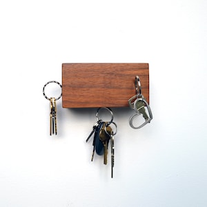May include: A wooden key holder with a dark brown finish. The key holder has three sets of keys hanging from it. One set of keys is silver and has a heart-shaped ring. The other two sets of keys are gold and have round rings.