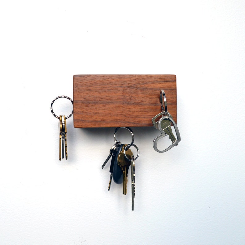 May include: A wooden key holder with a dark brown finish. The key holder has three sets of keys hanging from it. One set of keys is silver and has a heart-shaped ring. The other two sets of keys are gold and have round rings.