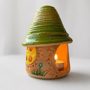 Può includere: Un portacandele a forma di fungo in ceramica con un tetto verde strutturato e una base marrone. La base presenta una finestra gialla, decorazioni floreali e una candela accesa all'interno, creando un bagliore caldo.
