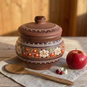 May include: A brown clay pot with a matching lid, featuring hand-painted white flowers and orange fruit motifs. The pot is accompanied by a wooden spoon, a red apple, and small white flowers, all arranged on a light-colored fabric napkin.