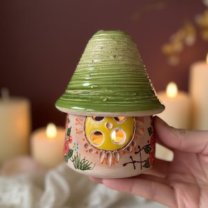 Op de afbeelding: Een keramische paddenstoelvormige kandelaar met een groene, getextureerde bovenkant en een bloemversierde basis. De kandelaar heeft een gele raam en wordt in een hand gehouden. Kaarsen en bloemblaadjes staan op de achtergrond.