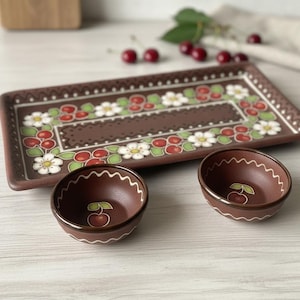 Puede incluir: Una bandeja de cerámica marrón y dos cuencos pequeños con un diseño de cerezas y flores. La bandeja es rectangular con un borde decorativo y los cuencos tienen un borde ondulado. Hay cerezas frescas esparcidas alrededor de la bandeja.