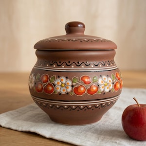 May include: A brown ceramic jar with a lid, adorned with a floral pattern of white flowers and red fruit. The jar has a rounded shape and is decorated with black and white wavy lines. A red apple rests on a white cloth nearby.