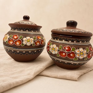 Könnte beinhalten: Zwei braune Keramikgläser mit Deckel. Die Gläser sind mit weißen Blumen, roten Früchten und grünen Blättern verziert. Die Gläser haben einen dunkelbraunen Boden und Deckel mit einem kleinen Knopf oben. Die Gläser stehen auf einem beigen Tuch.