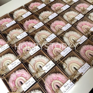 May include: A display of rainbow-shaped decorations, each in a small, square cardboard box. The rainbows are in shades of pink, white, and beige, with a fluffy texture. Each box is tied with twine and has a small tag attached.