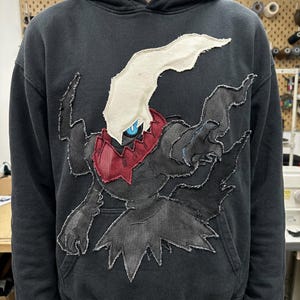 May include: A black hooded sweatshirt featuring a large, raw-edge fabric appliqué of a dark, shadowy creature with a cream-colored head crest, a red collar, and a single glowing blue eye, stitched onto the center of the chest.