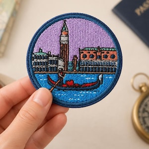 Peut inclure: Un écusson brodé circulaire représentant une scène de Venise, en Italie. Le motif comprend un haut clocher, des bâtiments historiques et une gondole rouge sur une eau bleue. L'écusson a une bordure cousue bleu foncé sur un ciel violet clair.