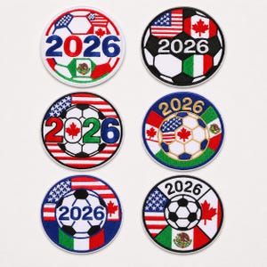 Pode incluir: Seis emblemas bordados circulares com designs de bolas de futebol, o ano 2026 e bandeiras nacionais dos Estados Unidos, Canadá e México. Cada emblema exibe um arranjo único destes elementos nas cores vermelho, branco, azul e verde.