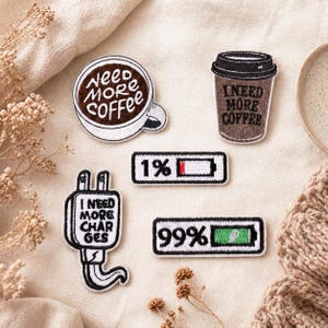 May include: A collection of embroidered patches with coffee and battery themes. One patch says "Need More Coffee" in a coffee cup, another on a to-go cup. Other patches show a battery at 1% and 99%, and a charger that says "I Need More Charges."