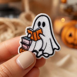 May include: An embroidered patch featuring a white ghost with black outlines holding an open orange book. The ghost sits beside a small stack of three books in shades of blue, pink, and orange. The design is rendered in white thread with black details.