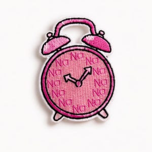 May include: A pink embroidered patch in the shape of an alarm clock. The clock face has the word "No" repeated multiple times. The hands of the clock point to the time of 10:10. The patch has a white border.