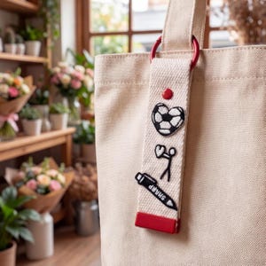 Puede incluir: Un bolso de tela beige con una correa vertical sujeta por una anilla roja. La correa muestra tres parches bordados: un balón de fútbol blanco y negro, una figura de palo sosteniendo un corazón y un marcador negro con la etiqueta SHARP.