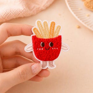 以下が含まれることがあります： 笑顔の漫画のフライドポテト容器の形をした赤い刺繍パッチ。 パッチには笑顔の顔、腕、足があります。 フライドポテトは金色です。
