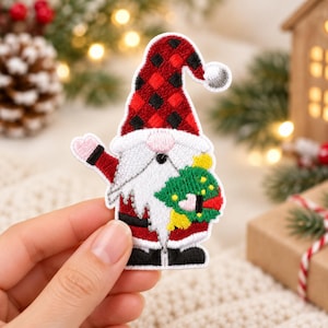 May include: An embroidered patch of a gnome wearing a red and black buffalo plaid hat with a white pom-pom. The gnome has a white beard, a pink nose, and is holding a green wreath with yellow accents. The gnome wears a red coat and black boots.