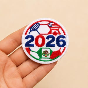 May include: A circular embroidered patch featuring a football design with the text 2026 in blue and red. The ball panels display the flags of the United States, Canada, and Mexico, incorporating red, white, blue, and green thread colours.