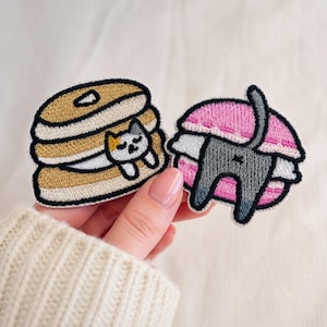 May include: Two embroidered patches. One patch features a stack of golden-brown pancakes with a small calico cat resting against them. The second patch depicts a gray cat tail and hind legs emerging from a pink and white sandwich cookie.