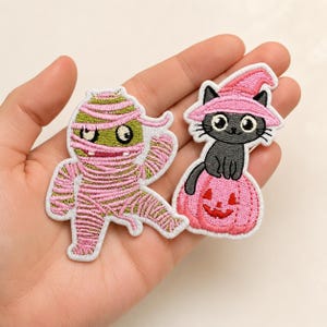 May include: Two embroidered patches featuring Halloween themes. One patch depicts a mummy wrapped in pink bandages with green skin and large eyes. The second patch shows a black cat wearing a pink witch hat, sitting atop a pink jack-o'-lantern.