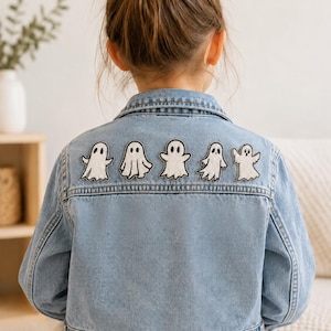 Puede incluir: Una chaqueta de mezclilla azul claro con cinco parches bordados de fantasmas blancos con contornos y rasgos faciales negros, dispuestos en una fila horizontal en la parte superior de la espalda, con cuello clásico y botones de metal.