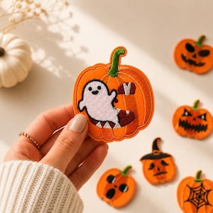 May include: An embroidered patch featuring an orange pumpkin with a green stem and a white ghost character inside. The ghost has black eyes and a small smile. The patch is held up, while other pumpkin patches with various faces are visible nearby.