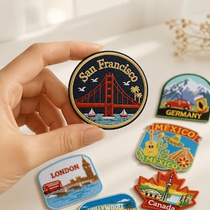 Può includere: Una toppa ricamata circolare raffigurante il Golden Gate Bridge in rosso, con il testo San Francisco in filo dorato su sfondo blu scuro, circondato da gabbiani bianchi e palme, con una base d'acqua azzurra.