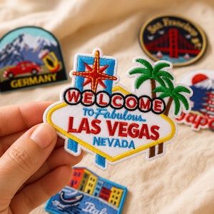 Op de afbeelding: Een geborduurde patch in de vorm van het Welcome to Fabulous Las Vegas Nevada bord, met een rode ster, groene palmbomen en rode en blauwe tekst op een witte achtergrond met een gele rand, omringd door andere patches met reisthema.