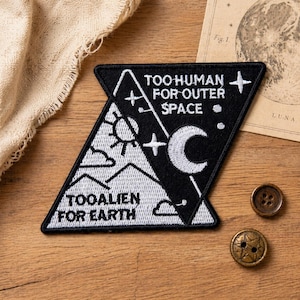 May include: A black and white embroidered patch with the text "TOO HUMAN FOR OUTER SPACE" and "TOO ALIEN FOR EARTH". The patch features a sun, moon, stars, and mountain design. Two decorative buttons and a vintage illustration are also visible.