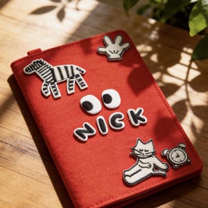 Könnte beinhalten: Ein roter Filz-Notizbuchumschlag mit aufgestickten Aufnähern. Zu den Aufnähern gehören ein Zebra, eine Cartoon-Hand, der Name "NICK", eine Katze und eine Uhr. Das Notizbuch ist geschlossen und hat eine kleine Lasche auf der linken Seite.