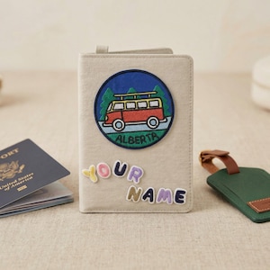 Könnte beinhalten: Ein beigefarbener Reisepasshalter mit einem farbenfrohen, gestickten Aufnäher mit einem roten Wohnmobil und dem Wort "ALBERTA". Der Halter hat die Worte "YOUR NAME" in bunten Buchstaben. Ein Reisepass und ein grünes Gepäckanhänger sind ebenfalls sichtbar.