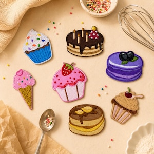 May include: A collection of embroidered patches featuring various desserts. Includes a cupcake with sprinkles, a chocolate cake with candles, a pink cupcake with a strawberry, a purple dessert with blueberries, a cupcake with a cherry, a pink ice cream cone, and pancakes.