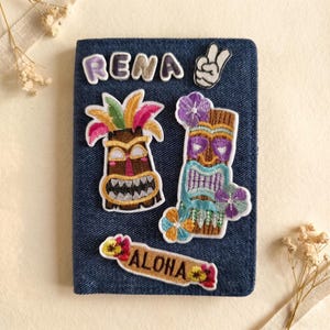 May include: A denim blue passport holder adorned with embroidered patches. Patches include the name "RENA", a peace sign, and two tiki-style figures. A patch with the word "ALOHA" is also present. The patches feature bright colours.
