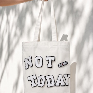 May include: A white tote bag with the words "NOT TODAY" in large, black-outlined letters. A small patch showing a battery at 1% is also on the bag. The bag is held by a hand against a white wall.