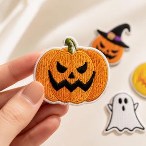Pode incluir: Um emblema bordado apresentando uma abóbora laranja com um caule verde e olhos e boca pretos em ziguezague. Outros emblemas são visíveis ao fundo, incluindo uma abóbora com um chapéu de bruxa e uma figura de fantasma branca.
